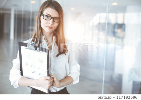 Beautiful female specialist with clipboard standing in sunny office and smiling charmingly. Business people concept 117192696