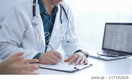 Doctor and a patient. The physician, wearing a white medical coat over a green shirt, is filling out a medical record form during a consultation in the clinic. Medical service 117192826