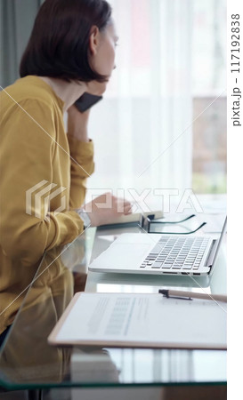 Young professional woman is talking by smartphone in modern office. Yellow blouse is passing to adult businesswoman. Business people concept 117192838