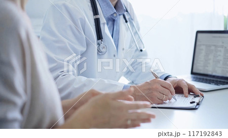 A doctor and a patient. The physician, wearing a white medical coat over a blue shirt, is filling out a medical record form during a consultation in the clinic. Medical service A doctor and a patient. The physician, wearing a white medical coat over a blue shirt, is filling out a medical record form during a consultation in the clinic. Medical service 117192843