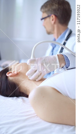Doctor man wearing white protective gloves is using ultrasound equipment on the neck of patient woman, lying for examination. Medicine concept 117193022