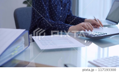 A female accountant with blue dotted blousy is using a laptop computer and calculator to calculate taxes at a glass desk in the office A female accountant with blue dotted blousy is using a laptop computer and calculator to calculate taxes at a glass desk in the office 117193300