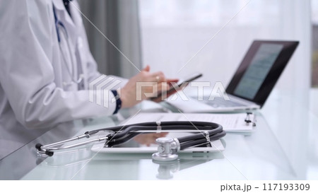 Stethoscope and clipboard are lying on the glass table while doctor woman is using a smartphone at the background. Medicine concept 117193309