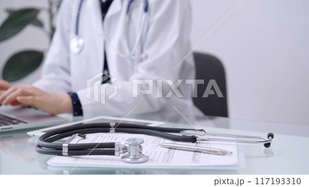 Clipboard with medical records and stethoscope are lying on the glass table while doctor woman is using tablet computer on the glass table. Medicine concept Clipboard with medical records and stethoscope are lying on the glass table while doctor woman is using tablet computer on the glass table. Medicine concept 117193310