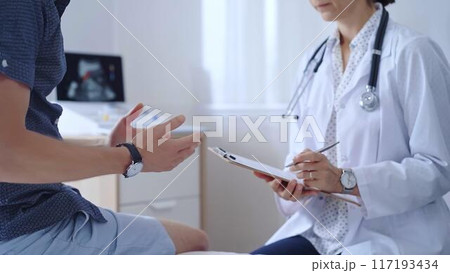 Doctor woman is taking notes on a clipboard while a male patient describes his symptoms in clinic procedure cabinet. Medicine service 117193434
