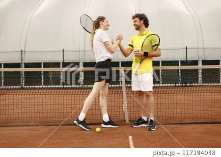 Curly man and woman tennis players standing on open summer court with rackets and balls in hands. Couple friendly shaking hands at court before the match. Man wearing yellow t-shirt and woman the Curly man and woman tennis players standing on open summer court with rackets and balls in hands. Couple friendly shaking hands at court before the match. Man wearing yellow t-shirt and woman the 117194038