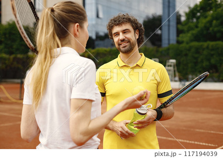 Curly man and woman tennis players standing on open summer court with rackets and balls in hands. Couple friendly chatting before the match. Man wearing yellow t-shirt and holding a bottle with water. 117194039