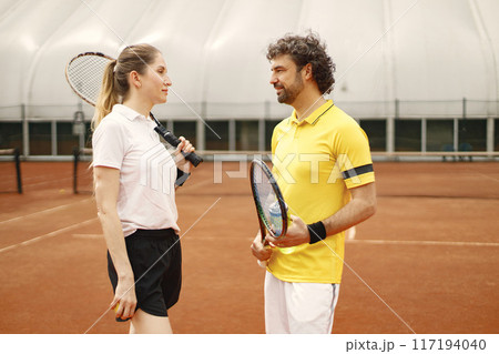 Curly man and woman tennis players standing on open summer court with rackets and balls in hands. Couple friendly chatting bedore the match. Man wearing yellow t-shirt and woman white one. Curly man and woman tennis players standing on open summer court with rackets and balls in hands. Couple friendly chatting bedore the match. Man wearing yellow t-shirt and woman white one. 117194040