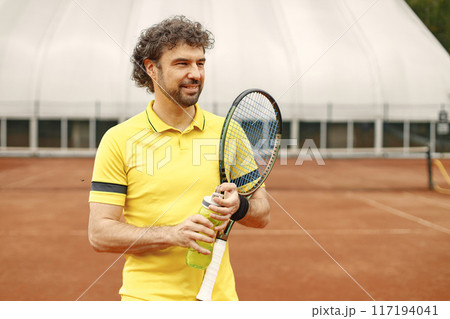 Portrait of tennis player man, front view. Man standing on a court and holding a racquet. Man wearing yellow t-shirt. 117194041