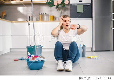 Portrait of a woman sitting in a kitchen on a floor and wearing red gloves. Caucasian woman after doing housework. Adult woman looking on her watch and have surprised face. Portrait of a woman sitting in a kitchen on a floor and wearing red gloves. Caucasian woman after doing housework. Adult woman looking on her watch and have surprised face. 117194043