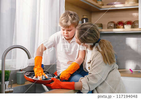 Blonde boy and adult woman cleaning in a kitchen and wearing orange gloves. Caucasian boy washing a frying pan with detergents. Mother supporting her son. Blonde boy and adult woman cleaning in a kitchen and wearing orange gloves. Caucasian boy washing a frying pan with detergents. Mother supporting her son. 117194048
