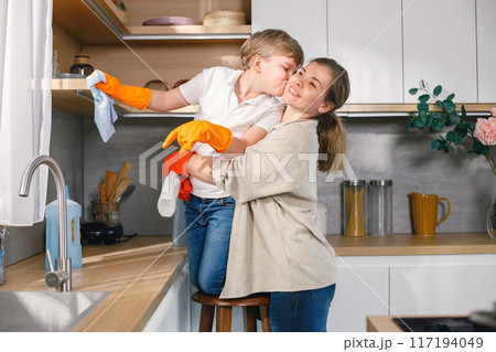 Blonde boy and adult woman cleaning in a kitchen and wearing orange gloves. Caucasian boy helping his mother to do housework. Mother hugging her son. Blonde boy and adult woman cleaning in a kitchen and wearing orange gloves. Caucasian boy helping his mother to do housework. Mother hugging her son. 117194049