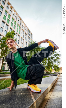 Young man, breakdancer in green shirt and black jacket executes stylish pose on ledge, showcasing balance and flair. 117195529