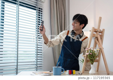 Young Artist Taking a Selfie in Studio with Easel and Art Supplies, Wearing Apron and Headphones Young Artist Taking a Selfie in Studio with Easel and Art Supplies, Wearing Apron and Headphones 117195565