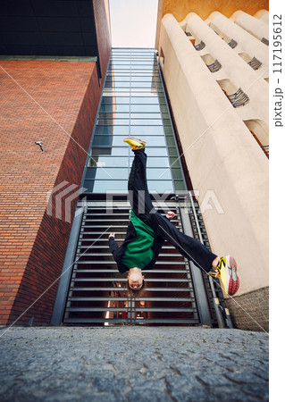 Breakdancer flips upside down, his bright yellow sneakers contrasting against towering buildings and creating dynamic visual. Breakdancer flips upside down, his bright yellow sneakers contrasting against towering buildings and creating dynamic visual. 117195612