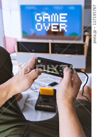 Male hands hold joystick video-game console next blue TV-screen with text Game Over. First-person view. First-person view. Home entertainment concept Male hands hold joystick video-game console next blue TV-screen with text Game Over. First-person view. First-person view. Home entertainment concept 117195767
