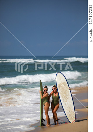 Two beautiful blondes in swimsuits pose with surfboards on the beach Two beautiful blondes in swimsuits pose with surfboards on the beach 117197253