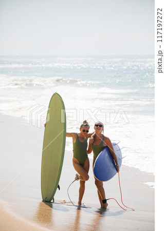Two beautiful blondes in swimsuits pose with surfboards on the beach Two beautiful blondes in swimsuits pose with surfboards on the beach 117197272