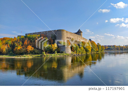 Scenic view of Ivangorod Fortress surrounded by colorful autumn trees, reflecting in calm river below under clear blue sky Scenic view of Ivangorod Fortress surrounded by colorful autumn trees, reflecting in calm river below under clear blue sky 117197901