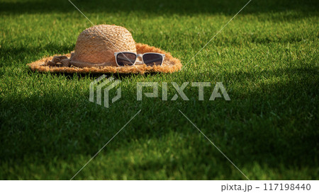 A straw hat adorned with white sunglasses sits on vibrant green grass, with purple flowers blooming in the background, symbolizing a peaceful summer afternoon. 117198440