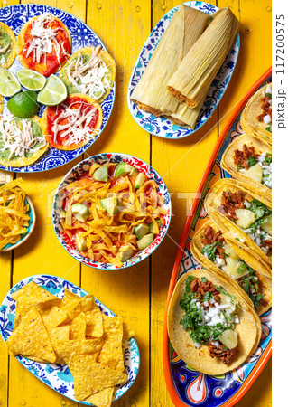 Assortment of colorful Mexican dishes on a vibrant yellow table, featuring tacos, tamales, aztek soup, totopos Assortment of colorful Mexican dishes on a vibrant yellow table, featuring tacos, tamales, aztek soup, totopos 117200575