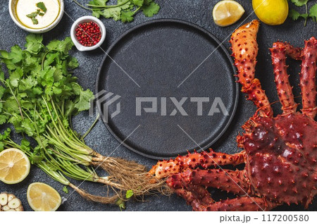 Top view of fresh king crab legs, herbs, and lemon wedges surrounding an empty black plate on a dark textured background, ready for a gourmet seafood preparation 117200580