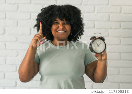 Surprised african american woman holding alarm clock amazed with open mouth and surprise happy face at home, on brick background. Being late and time passing quickly concept Surprised african american woman holding alarm clock amazed with open mouth and surprise happy face at home, on brick background. Being late and time passing quickly concept 117200818