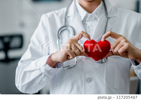 A healthcare professional in a white coat holding a red heart, symbolizing care, compassion, and health in a medical environment. A healthcare professional in a white coat holding a red heart, symbolizing care, compassion, and health in a medical environment. 117205487