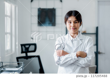 A confident female doctor wearing a white coat and holding a stethoscope, standing in a modern medical office setting. 117205488