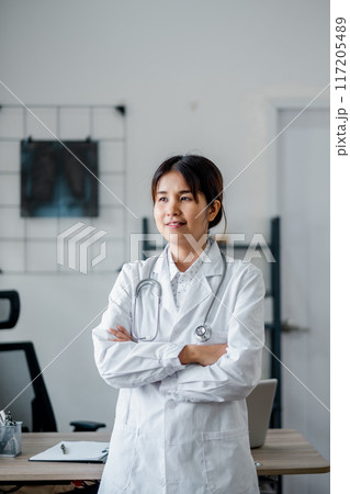 A confident female doctor in a white coat with a stethoscope stands in a modern medical office, ready to assist patients. A confident female doctor in a white coat with a stethoscope stands in a modern medical office, ready to assist patients. 117205489