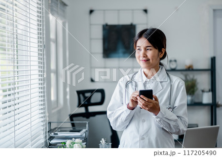 A young female doctor in a white coat using a smartphone in a modern medical office with bright natural light coming through the window. A young female doctor in a white coat using a smartphone in a modern medical office with bright natural light coming through the window. 117205490