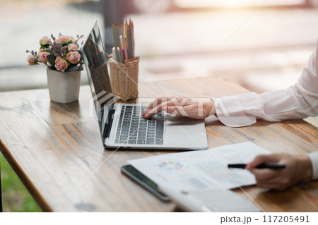A businesswoman's hand typing on a laptop at a wooden desk with documents, flowers, and office supplies in a well-lit office environment. A businesswoman's hand typing on a laptop at a wooden desk with documents, flowers, and office supplies in a well-lit office environment. 117205491