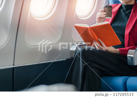 A businesswoman in a red blazer reads a book and drinks coffee while seated on an airplane during a flight. A businesswoman in a red blazer reads a book and drinks coffee while seated on an airplane during a flight. 117205513