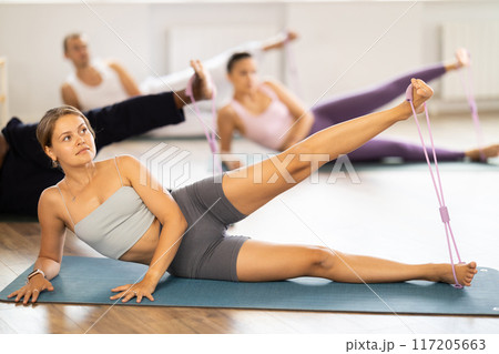 Pilates class - group of people of different nationalities doing leg stretches using resistance bands in gym 117205663