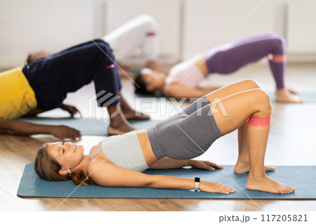 Girl performs glute bridge during Pilates multinational training with elastic band on shins 117205821