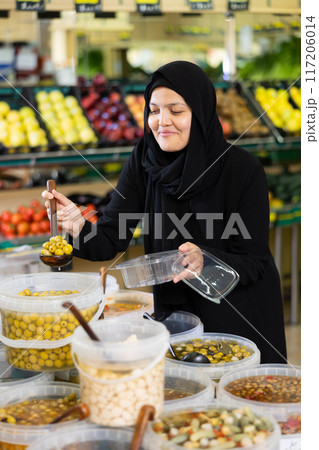 Asian girl in hijab buys pickled olives by weight in supermarket 117206014