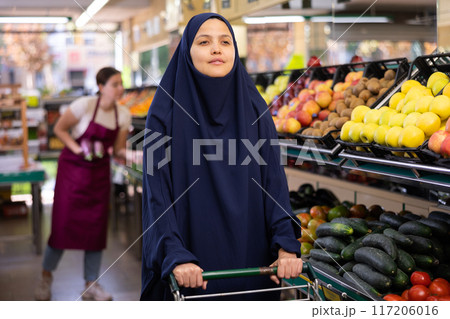 Muslim woman in dark blue hijab stands in grocery department of supermarket with grocery basket 117206016