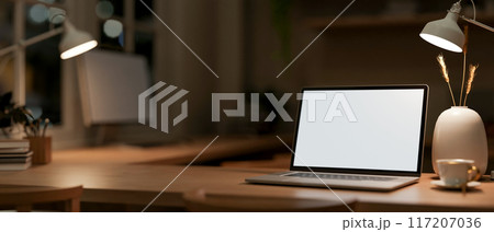A laptop computer mockup on a wooden table in an office at night, illuminated by table lamps. 117207036