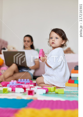 cute little baby girl playing with toys and mom sitting on floor with laptop in room cute little baby girl playing with toys and mom sitting on floor with laptop in room 117210301