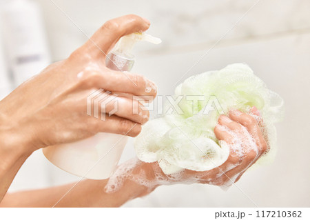 female hands applying liquid soap shower gel from white cosmetic bottle on sponge in bathroom 117210362