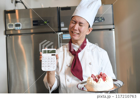 厨房で電卓とケーキを持ち悩む男性料理人 117210763