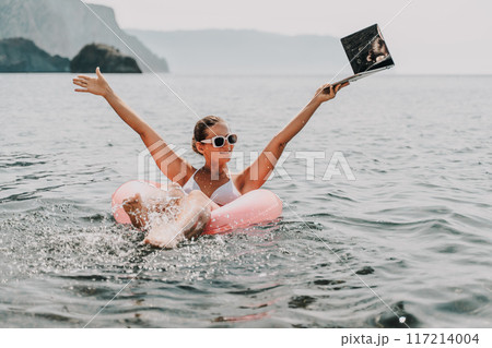 A woman is floating in a pink inflatable raft with a laptop in her hand. She is smiling and she is enjoying her time in the water. 117214004