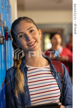 Smiling teenage girl leaning against lockers in high school hallway, holding tablet Smiling teenage girl leaning against lockers in high school hallway, holding tablet 117214349