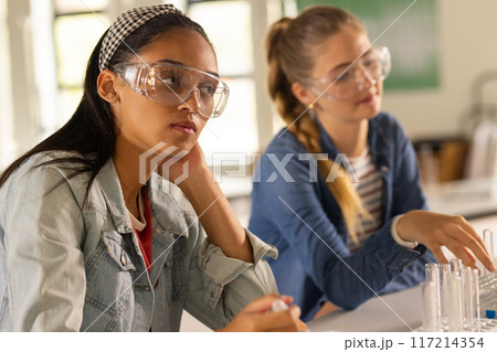 Wearing safety goggles, teenage girls conducting science experiment in high school classroom 117214354