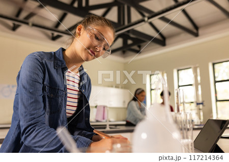 In high school, teenage girl writing notes in science classroom, wearing safety goggles In high school, teenage girl writing notes in science classroom, wearing safety goggles 117214364
