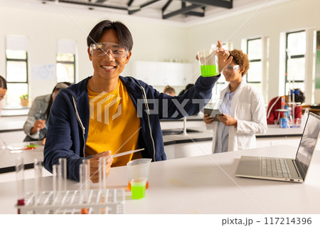 In high school laboratory, teenager holding beaker with green liquid, smiling at camera 117214396