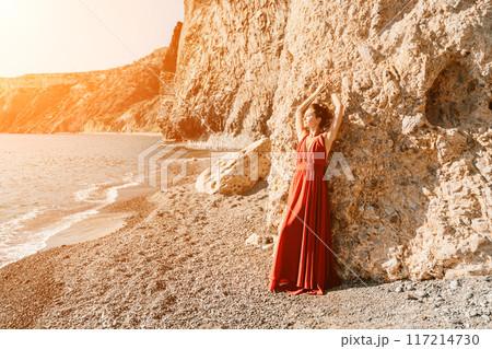 Woman red dress sea. Woman in a long red dress posing on a beach with rocks on sunny day. Girl on the nature on blue sky background. Woman red dress sea. Woman in a long red dress posing on a beach with rocks on sunny day. Girl on the nature on blue sky background. 117214730