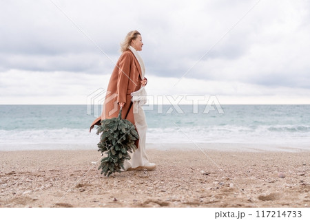 Blond woman Christmas tree sea. Christmas portrait of a happy woman walking along the beach and holding a Christmas tree in her hands. She is wearing a brown coat and a white suit. Blond woman Christmas tree sea. Christmas portrait of a happy woman walking along the beach and holding a Christmas tree in her hands. She is wearing a brown coat and a white suit. 117214733