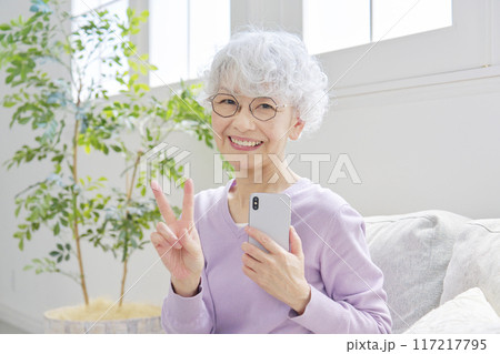 スマートフォンを持ってピースサインするシニア女性 リビングルーム スマートフォンを持ってピースサインするシニア女性 リビングルーム 117217795