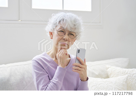 スマートフォンを持って悩むシニア女性　リビングルーム　カメラ目線 117217798